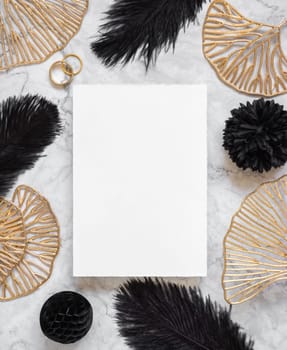 Blank card on marble table near black feathers and golden leaves with wedding rings top view. Greeting or Invitation card mockup. Elegant femminine flat lay with vertical paper card