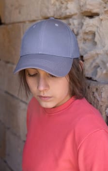 Hipster Teen girl in grey baseball cap and pink long sleeve shirt looking down. Cap and shirt mockup. Serious Teenager girl near a stone wall outdoor. Loneliness concept