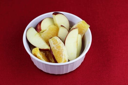 White breakfast fruit of red apple and orange sections on a red place mat close-up. Breakfast food fruit slices of orange citrus and red apple food in a round white bowl on a red background placemat.