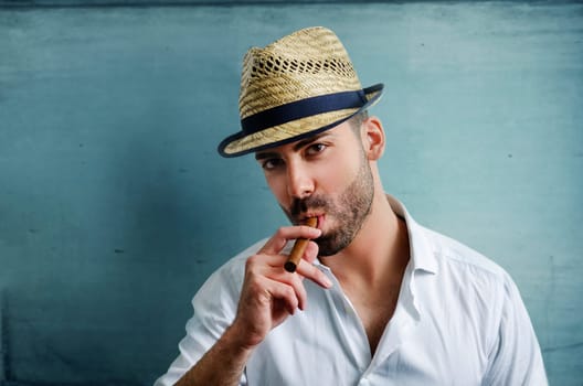 Young man with modern hat smoking a cuban cigar in front of blue wall