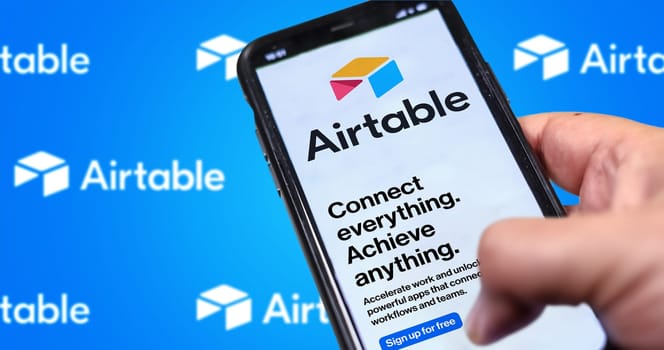 San Francisco, CA, US, May 2022: Male hand holding a phone with Airtable company web page on screen. In the background the logo Airtable blurred and repeated. Airtable is a cloud collaboration service