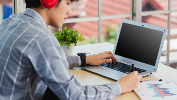 Asian businessman writing notes with pen inspired ideas list on paper notepad while working on laptop computer. Happy handsome man sitting busy on desk table working from home office doing note