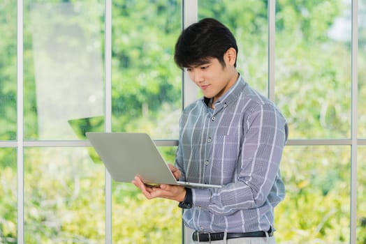 Asian businessman smiling and working on a laptop computer stand near window in office. Portrait of handsome man standing and online meeting with computer notebook in office lobby