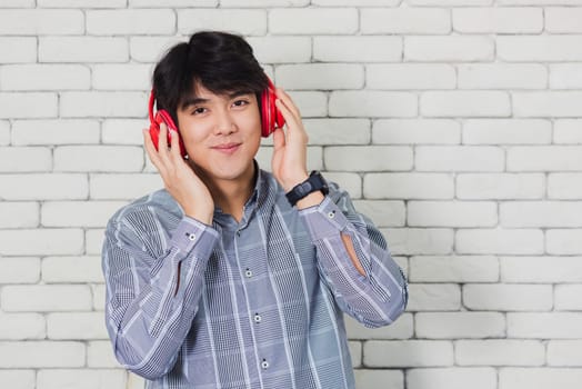 Portrait of Asian handsome young man he smile listening to music in headphones on brick wall background. Happy teenage guy smiling. Happy laughing man standing with red headphones