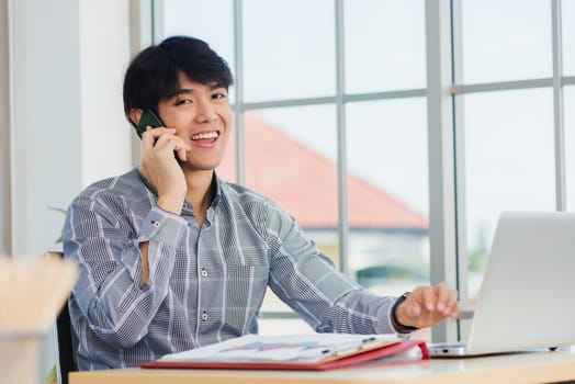 Asian businessman smiling and talking on a mobile phone at home office desk. The man smile call on smartphone with customer at the house, making sell offer