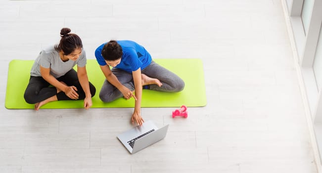 Overhead two Asian lifestyle adult and young woman in sportswear sitting floor on mat with dumbbells look laptop computer for training videos online in yoga studio, fit sport healthy workout exercise