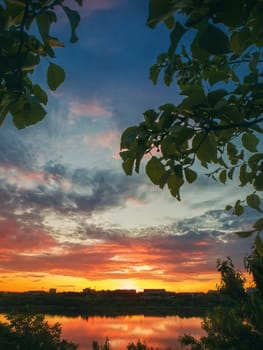 Gorgeous summer sunset as seen from the garden among the green trees. Sundown view over the city horizon reflected on the calm Delia lake water in Ungheni city, Moldova