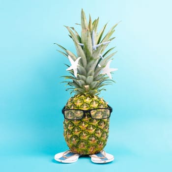 Pineapple with glasses and flip flops, vacation and travel concept, tropical summer holiday, funny fruit, blue background