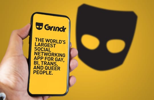San Francisco, CA, US, May 2022: hand holding a phone with Grindr mobile application on screen. In background the Grindr logo blurred. Grindr is dating application for gay, bi, trans, and queer people