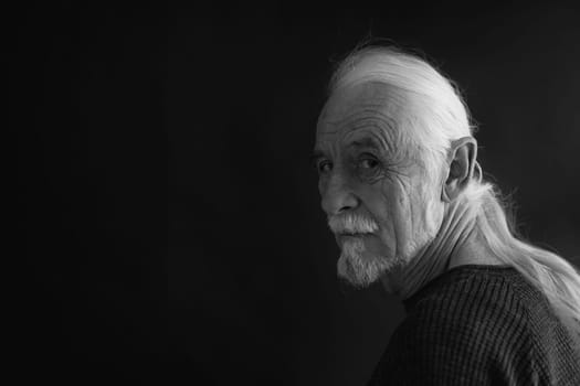 Low key studio black and white portrait of beautiful old man with gray hair in a braid looking over his shoulder at the camera. Horizontally.