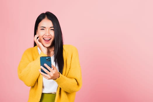 Happy Asian portrait beautiful cute young woman teen smiling excited using smart mobile phone studio shot isolated on pink background, Thai female surprised making winner gesture on smartphone