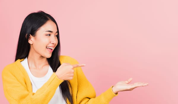Portrait Asian beautiful young woman smiling standing pointing finger out on pink background, Thai happy face excited female point into empty looking to side away with copy space for text