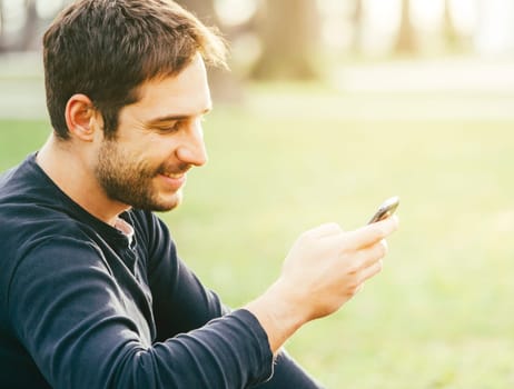Young happy man sitting in the park and using smartphone.