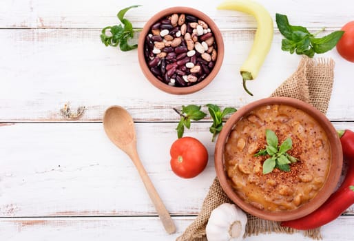 Traditional homemade recipe for spicy baked beans cooked in vintage clay bowl with vegetable and herbs. Top view of prepared dish and ingredients on white wooden table with copyspace