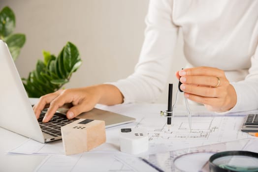 Architectural project workplace. Architect typing laptop keyboard to review design of house building before editing or sketching with compass on blueprint paper on table desk at architecture office