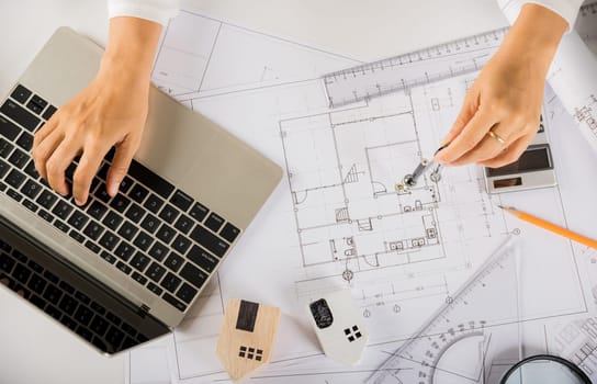 Architectural project workplace. Architect typing laptop keyboard to review design of house building before editing or sketching with compass on blueprint paper on table desk at architecture office