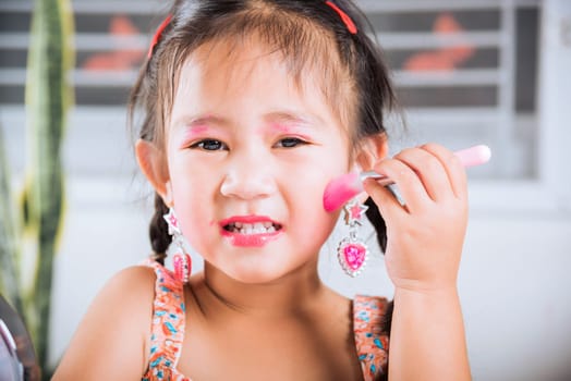 Asian adorable funny little girl making makeup her face she look in the mirror and cheek fluffy brush for powder, Learning activity to be woman, happy kid is beautiful make up with cosmetics toy