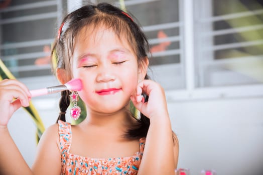 Asian adorable funny little girl making makeup her face she look in the mirror and cheek fluffy brush for powder, Learning activity to be woman, happy kid is beautiful make up with cosmetics toy