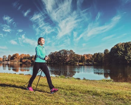 Vintage retro effect filtered hipster style image of nordic walking adventure and exercising - young woman hiking with nordic walking poles in park along river