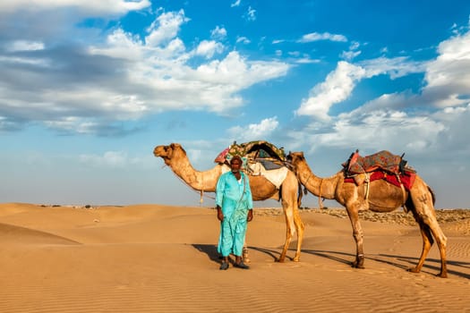 Rajasthan travel background - Indian cameleer (camel driver) with camels in dunes of Thar desert. Jaisalmer, Rajasthan, India