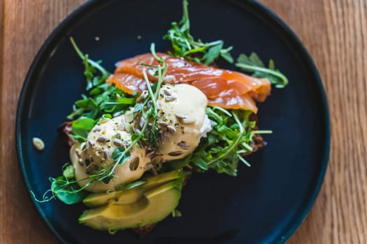 Egg benedict with arugula, salmon and avocado on a dark plate in the restaurant for breakfast.