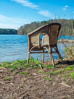 Wicker chair on the shore of the Lake Flaken near Wolterddorf in Brandenburg in a blue sky with veil clouds in April 2022 in a vertical orientation.