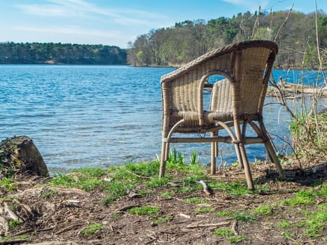 Wicker chair on the shore of the Flakensee near Wolterddorf in Brandenburg in a blue sky with veil clouds in April 2022.