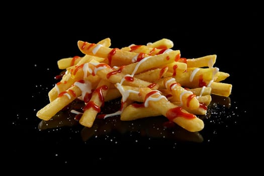 Delicious standard cut French fries topped with ketchup and mayonnaise placed on surface with sprinkled salt against black background in studio