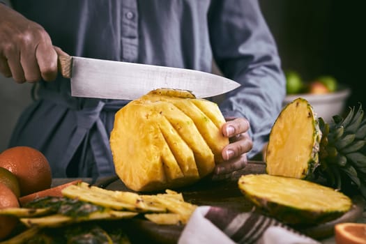 Unrecognizable female in blue shirt slicing ripe peeled pineapple while preparing food in kitchen at home