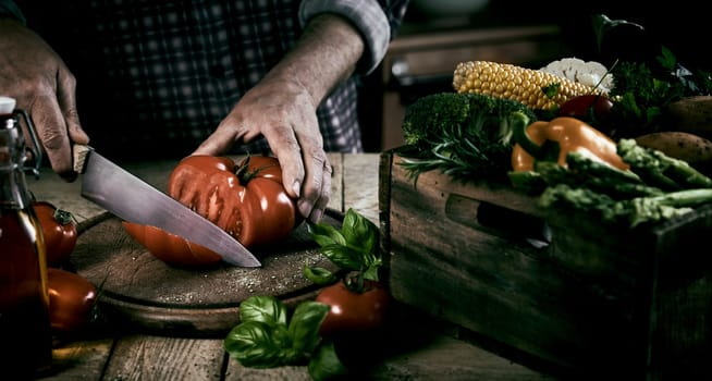 High angle of crop unrecognizable male chef cutting fresh ripe tomato on chopping board while standing at table with wooden box with assorted vegetables during dinner preparation