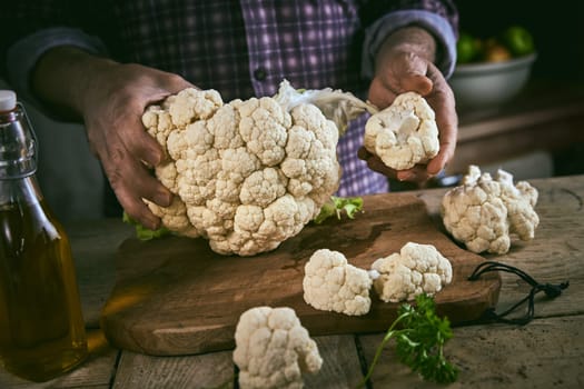 Anonymous male in checkered shirt dividing ripe cauliflower on smaller pieces on wooden cutting board near bottle of oil in rustic kitchen at home