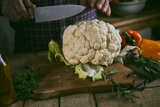Anonymous male with sharp knife preparing to cut whole cauliflower with leaves on lumber chopping board near rosemary and bell pepper in rustic kitchen at home
