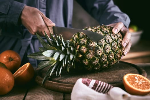 Unrecognizable woman in blue shirt using sharp knife to remove leaves from fresh pineapple near ripe oranges on table in kitchen at home