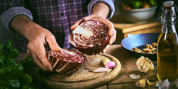 From above anonymous male in checkered shirt halving ripe red cabbage on round cutting board near cooking ingredients while preparing healthy dish in rustic kitchen