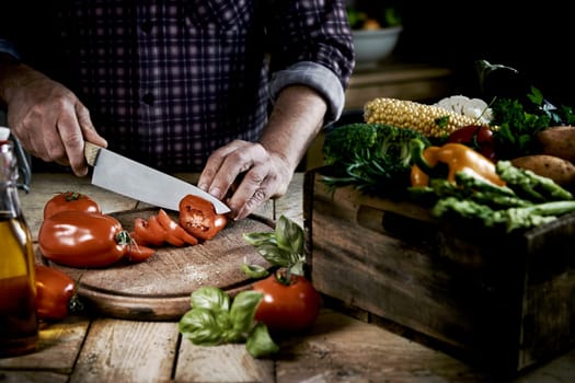From above of crop anonymous male chef in checkered shirt slicing fresh ripe tomatoes on round shaped wooden cutting board at table with box of assorted raw vegetables in kitchen