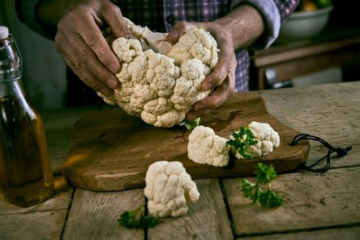 Crop man in checkered shirt tearing fresh cauliflower in pieces over shabby table with wooden cutting board and bottle of oil in rustic kitchen