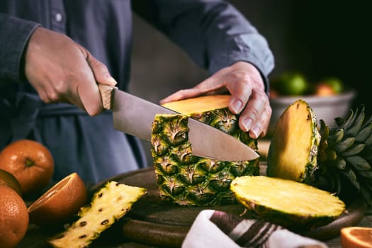Crop female in blue shirt removing peel from ripe pineapple near fresh oranges while preparing healthy food in kitchen at home