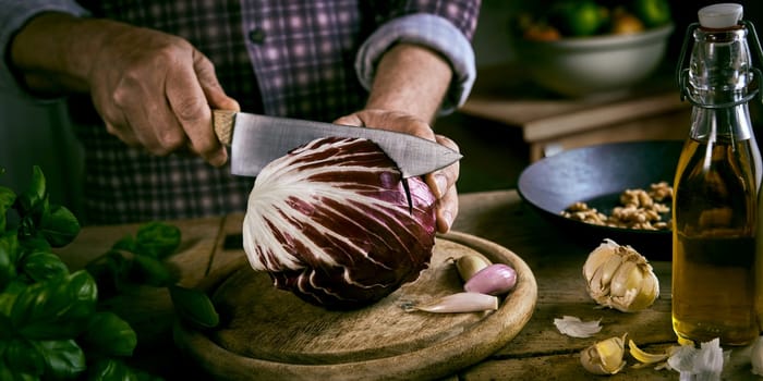 High angle of anonymous male in checkered shirt cutting fresh red cabbage in half with sharp knife near various ingredients while cooking healthy lunch in rustic kitchen at home