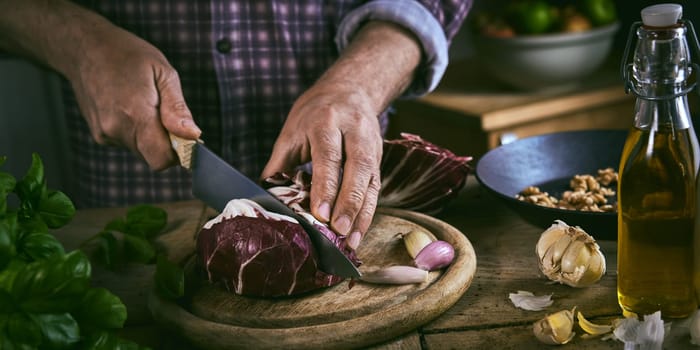 High angle of anonymous male in checkered shirt chopping ripe red cabbage with sharp knife near fresh cooking ingredient during lunch preparation at home