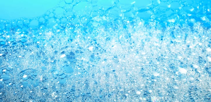 Background with fragile bubbles of white soap foam covering surface of bright blue water