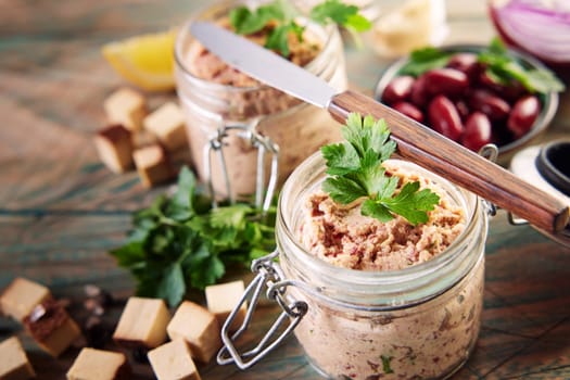 From above of yummy vegetarian liverwurst pate in glass jars decorated with leaves of fresh parsley served on wooden table near bowl of red beans and cubed cheese in kitchen