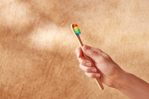 Crop unrecognizable female showing eco friendly bamboo toothbrush with colorful bristles against blurred beige background