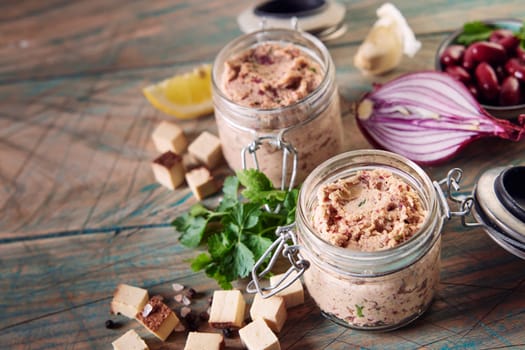 High angle of glass jars with appetizing homemade pate placed on wooden table near scattered cubed cheese herbs and halved onion in kitchen in daylight
