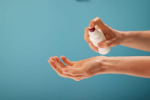Unrecognizable female disinfecting hands with sanitizer from plastic spray bottle against turquoise background