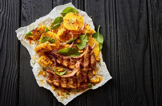 Top view of delectable grilled lamb chops with corn slices and green spinach leaves served on white parchment on black wooden background
