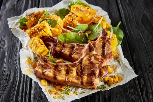 From above of yummy grilled lamb chops and slices of corn and orange served on white parchment with spinach on black wooden table