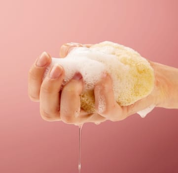 Womans hand squeezing water out of a natural sea sponge. Body care photography on a feminine pink background color.
