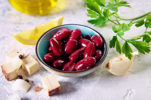 From above of delicious red kidney beans in small bowl served on table near cheese cubes garlic clove and lemon slice arranged with parsley and carafe of olive oil