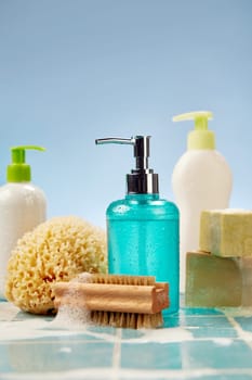 Foamy brush placed near sponge and soap on wet tiled shelf during hygienic routine against blue background