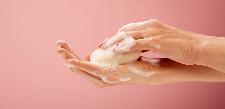 Young woman cleaning hands with foamy soap on a bright pink background. With a piece of white creamy soap for skin care projects